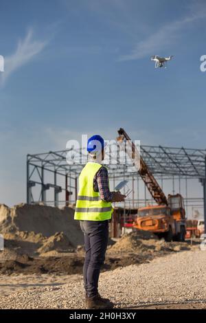 Rückansicht des Ingenieurs, der die Drohne über der Baustelle steuert, mit Metallkonstruktion und Kran im Hintergrund Stockfoto