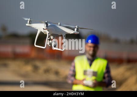 Nahaufnahme der Drohne, die auf der Baustelle fliegt, und des Bauarbeiters mit Fernbedienung im Hintergrund Stockfoto