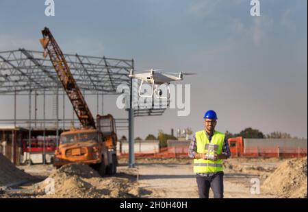 Glücklicher Ingenieur, der Drohne über der Baustelle mit Metallkonstruktion und Kran im Hintergrund steuert Stockfoto