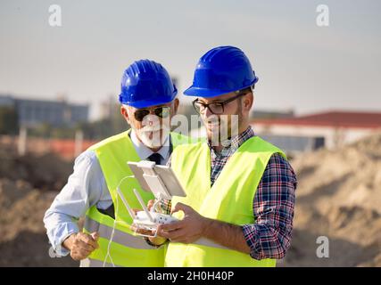 Zwei Ingenieure mit Helmen und Westen, die per Fernbedienung mit der Drohne arbeiten. Technologische Innovationen in der Bauindustrie Stockfoto