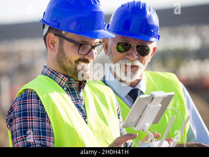 Zwei Ingenieure mit Helmen und Westen, die per Fernbedienung mit der Drohne arbeiten. Technologische Innovationen in der Bauindustrie Stockfoto