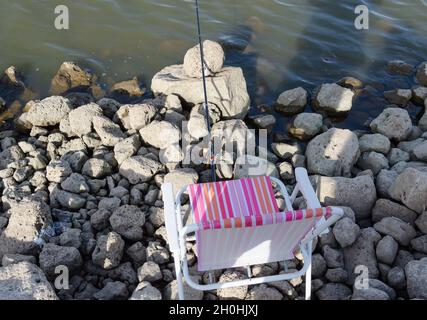 Am felsigen Ufer des Stausees befindet sich eine Angelrute mit einer Walze und einem Klappstuhl daneben Stockfoto