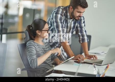 Junge, multirassische Kolleginnen und Kollegen, die auf den Laptop-Bildschirm schauen und über das Projekt diskutieren, arbeiten im modernen Coworking Space zusammen Stockfoto