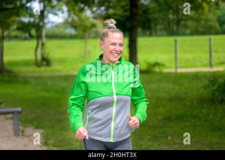 Glückliche gesunde Frau mit einem lebhaften Lächeln joggen in einem ländlichen Park an der Kamera vorbei mit einem strahlenden Lächeln voller Vitalität Stockfoto