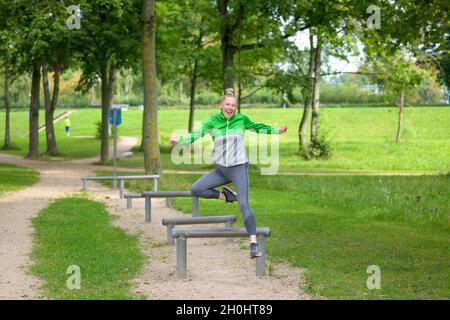 Glückliche, gesunde, funstige Frau, die in einem Park mit einem strahlenden Lächeln in einem aktiven Lifestyle-Konzept die Hürden beim Springen in der Luft auf die Kamera anrückt Stockfoto