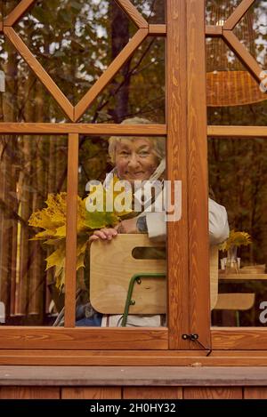 Das Foto wurde durch ein Fenster aufgenommen, in dem Bäume reflektiert werden. Eine ältere Frau sitzt am Fenster mit einem Strauß gelber Ahornblätter. Stockfoto