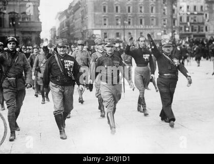 „Nachdem Mussolini am Morgen des 31. Oktober 1922 als Regierungschef vereidigt wurde, marschierten seine faschistischen Anhänger, die Blackshirts, am selben Tag in einer großen „Siegesparade“ durch Rom, um die „Marsch gegen Rom“-Propaganda zu vervollständigen. Das Bild zeigt die Squadristen, die auf die Piazza Venezia marschieren, angeführt von ehemaligen Generälen der italienischen Armee (erkennbar an den Greca auf ihrer Lagerkappe). Der zweite von links ist der Kriegsgeneral Luigi Capello, der wahrscheinlich nach rechts geht, ist General Armando Diaz (Stabschef im Ersten Weltkrieg). Sie kommen von der Via del Corso. Im Rücken an Stockfoto