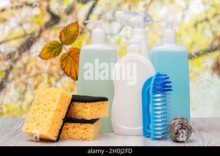 Flaschen mit Spülmittel und Schwämmen vor dem Fenster mit Wassertropfen. Stockfoto