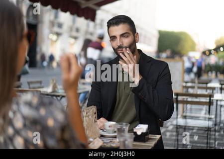 Junger Mann, der mit einer Frau, die Espresso trinkt und Zigaretten raucht, auf einem Tisch sitzt - Dating oder Geschäftskonzept. Stockfoto