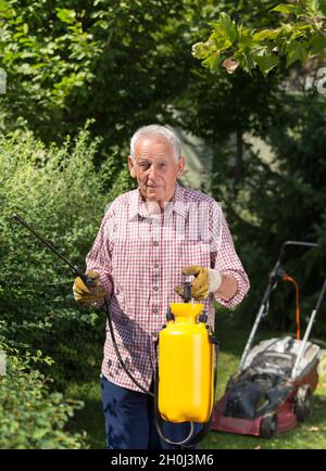 Senior Mann sprüht Hecke im Garten. Mäher im Hintergrund. Pflege der Pflanzen Konzept Stockfoto
