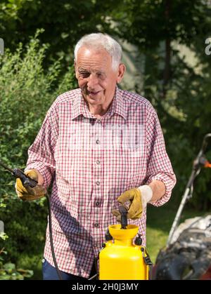 Senior Mann sprüht Hecke im Garten. Mäher im Hintergrund. Pflege der Pflanzen Konzept Stockfoto