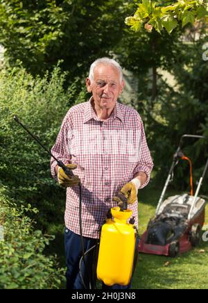 Senior Mann sprüht Hecke im Garten. Mäher im Hintergrund. Pflege der Pflanzen Konzept Stockfoto