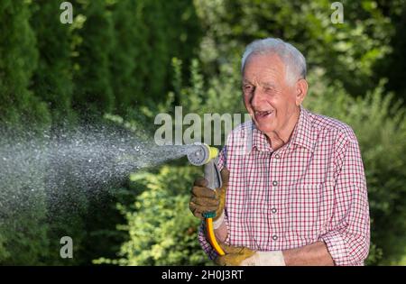 Glücklicher älterer Mann, der Pflanzen im Garten aus einer Handpistole auf einem Schlauch gießt Stockfoto