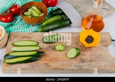 Seitenansicht von ganzen in Scheiben geschnittenen und geschnittenen Gurken und schwarzem Pfeffer auf dem Schneidebrett mit Tomaten geschmolzenem Öl Schüssel mit Gurkenscheiben und auslaufenden Gurken Stockfoto