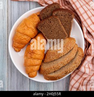 Draufsicht auf Brote, wie Croissant in Scheiben geschnittenes Roggen und gesäte braune Cola in Teller auf kariertem Tuch auf Holzhintergrund Stockfoto