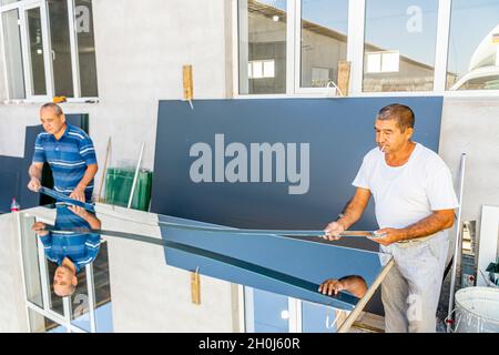 Zwei Arbeiter nehmen mit ihren Händen ein Stück geschnittenes Spiegelglas aus dem getrimmten Spiegelstück, um es in der lokalen Möbelproduktion zu verwenden Stockfoto