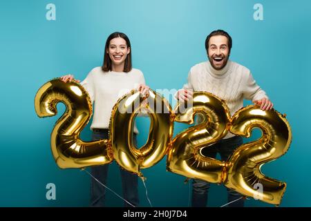 Fröhliches Paar, das die Kamera anschaut, während es goldene Ballons in Form von 2022 auf Blau hält Stockfoto