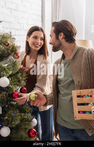 Fröhliches Paar lächelt sich gegenseitig an, während sie zu Hause Weihnachtsbaum dekorieren Stockfoto