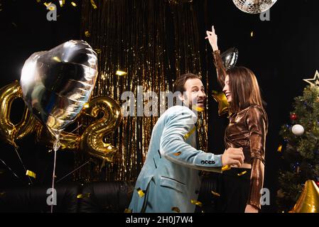 Fröhlicher Mann, der die Kamera anschaut, während er mit einer eleganten Frau während der Neujahr-Party auf Schwarz tanzt Stockfoto