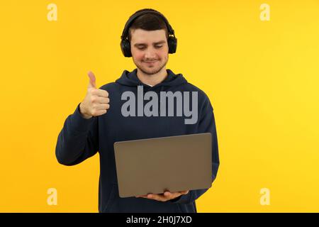 Ein junger Mann mit Kopfhörern hält den Laptop und zeigt die Daumen auf gelbem Hintergrund. Stockfoto