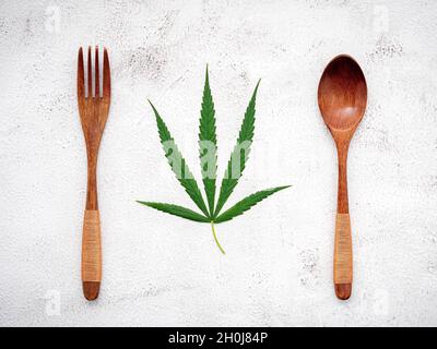 Essen konzeptuelles Bild eines Cannabisblattes mit Löffel und Gabel auf weißem Beton Hintergrund. Stockfoto