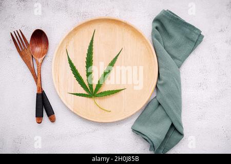 Essen konzeptuelles Bild eines Cannabisblattes mit Löffel und Gabel auf weißem Beton Hintergrund. Stockfoto