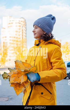 Ein lächelndes, niedliches Mädchen in einer hellen Jacke mit einem herbstlichen Bouquet aus leuchtend herbstlichen Blättern. Porträt eines glücklichen positiven Mädchens. Sonniger Herbsttag Hintergrund. S Stockfoto