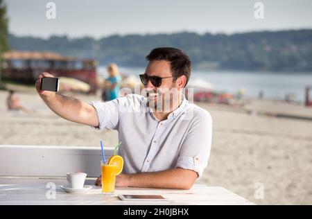 Ein hübscher Mann mit Sonnenbrille, der am Strand ein Handy zum Webchatten benutzt. Genießen Sie am Fluss Stockfoto