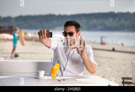 Ein hübscher Mann mit Sonnenbrille, der am Strand ein Handy zum Webchatten benutzt. Genießen Sie am Fluss Stockfoto