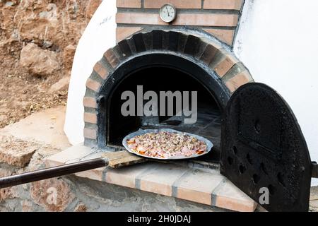 Mann Putting Pizza in handgefertigten weiß lackierten Holzofen im Freien mit Schaufel gebaut Stockfoto