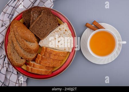 Draufsicht auf Brote, wie in Scheiben geschnittenes braunes Kob-Roggen-Baguette und Fladenbrot auf Plaid-Tuch auf dem Teller und eine Tasse heißer Toddy mit Zimt auf einer Untertasse auf Grau Stockfoto