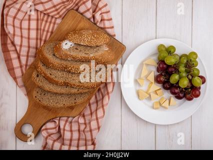 Draufsicht auf braun geschnittenes, gesätes Kohlenstück auf Schneidebrett auf kariertem Tuch und Teller mit Käse und Trauben auf Holzhintergrund Stockfoto