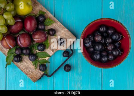 Draufsicht auf Früchte als Traubenpflüten mit Traubenbeeren und Blättern auf Schneidebrett und Schale mit Traubenbeeren auf blauem Hintergrund Stockfoto