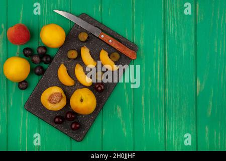 Draufsicht auf Früchte als ganze halb geschnittene Aprikosen und Schlehen-Beeren mit Messer auf Schneidebrett und auf grünem Hintergrund mit Kopierraum Stockfoto