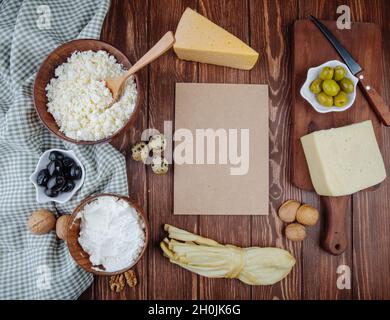 Draufsicht auf ein Stück braunes Papierblatt und Hüttenkäse in Holzschüsseln und verschiedenen Käse mit eingelegten Oliven, Wachteleiern und Walnüssen auf karierten Fal Stockfoto