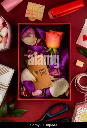 Draufsicht auf eine offene Geschenkbox mit korallenfarbener Rosenblüte und kleiner Papierkarte mit violettem Band und Blütenblättern auf rotem Hintergrund mit Schere, Hefter, Stockfoto