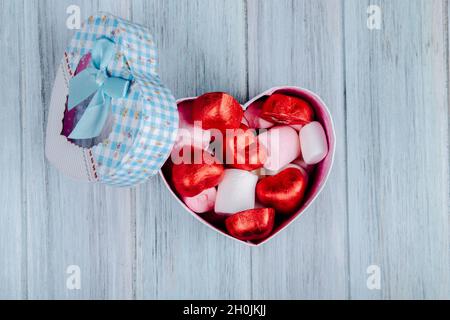 Draufsicht auf herzförmige Schokoladen-Bonbons in roter Folie mit rosa Marschmalin einer herzförmigen Geschenkbox auf grauem Holzhintergrund eingewickelt Stockfoto