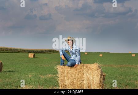 Hübscher Bauer mit Strohhut, der auf einem rollenden Ballen auf dem Feld sitzt Stockfoto