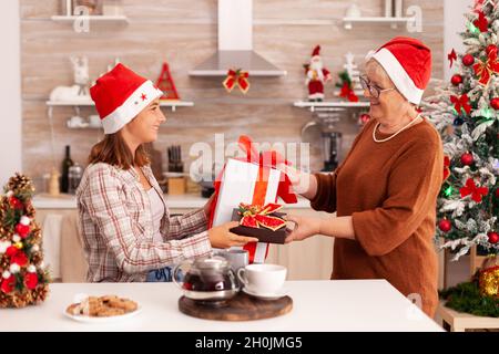 Großmutter Überraschende Enkelin mit Weihnachten Wrapper Geschenk feiert weihnachtsurlaub zusammen in dekorierter Küche. Kind genießt Wintersaison wechselnde Gegenwart mit Band auf sie mit gradma Stockfoto