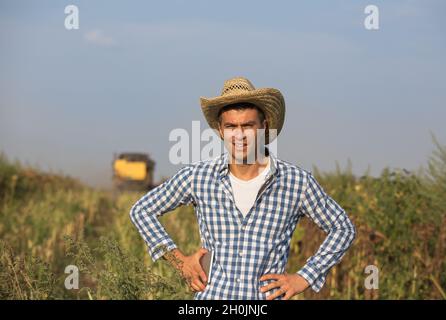 Hübscher Bauer mit Strohhut, der die Tablette hält und vor dem Mähdrescher im Sojabohnenfeld steht Stockfoto