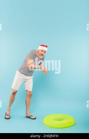 Die ganze Länge eines Mannes mittleren Alters in aufgesticktem T-Shirt und weihnachtsmütze imitiert das Tauchen in der Nähe des Schwimmrings auf Blau Stockfoto