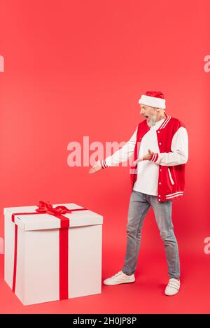 Die ganze Länge des schockierten Mannes mittleren Alters in santa hat zeigt auf riesige Gegenwart auf rot Stockfoto