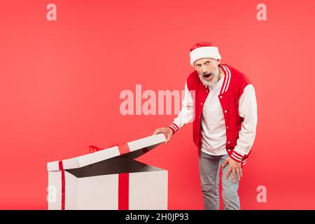 Schockiert Mann mittleren Alters in santa hat Eröffnung riesige Geschenk isoliert auf rot Stockfoto