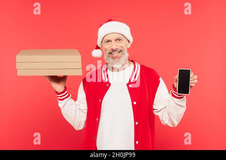Glücklicher Mann mittleren Alters in santa hat hält Pizzakisten und Smartphone mit leerem Bildschirm isoliert auf rot Stockfoto