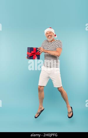 In voller Länge glücklich mittleren Alters Mann in santa Hut springen mit Geschenkbox auf blau Stockfoto