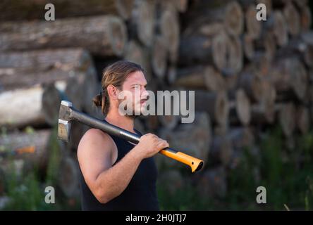 Junger, starker Holzfäller, der Axt auf der Schulter vor Holzstämmen auf dem Stapel trägt Stockfoto