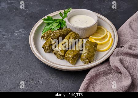 Traditionelles Gericht Dolma oder sarma, mit Petersilie, Kochtopf mit Sauerrahm und Zitrone in schwarzem Teller auf dunklem Holzhintergrund mit Leinenserviette Stockfoto