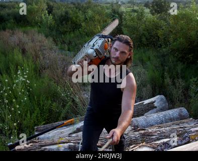 Ein junger, starker Holzfäller, der die Kettensäge auf der Schulter trägt und auf einen Haufen Baumstämme klettert Stockfoto