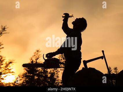 Silhouette von Holzfäller mit Kettensäge Trinkwasser aus der Flasche auf Holzstapel bei Sonnenuntergang in heißen Sommerabend Stockfoto