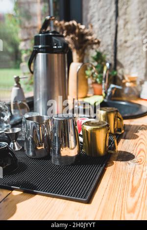 Kaffeemaschine, Thermoskanne und Metallutensilien auf einem Tisch am Waschbecken vor einem großen Küchenfenster mit hellem Tageslicht Stockfoto